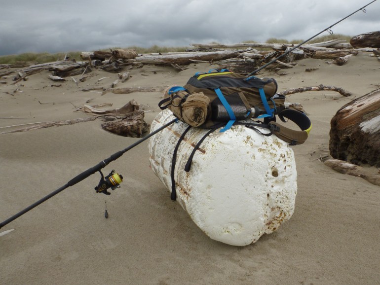 Fishing rod and backpack resting on a large cylindrical styrofoam float on the back beach. Backshore shelf with big wood in the background. Cloudy sky.