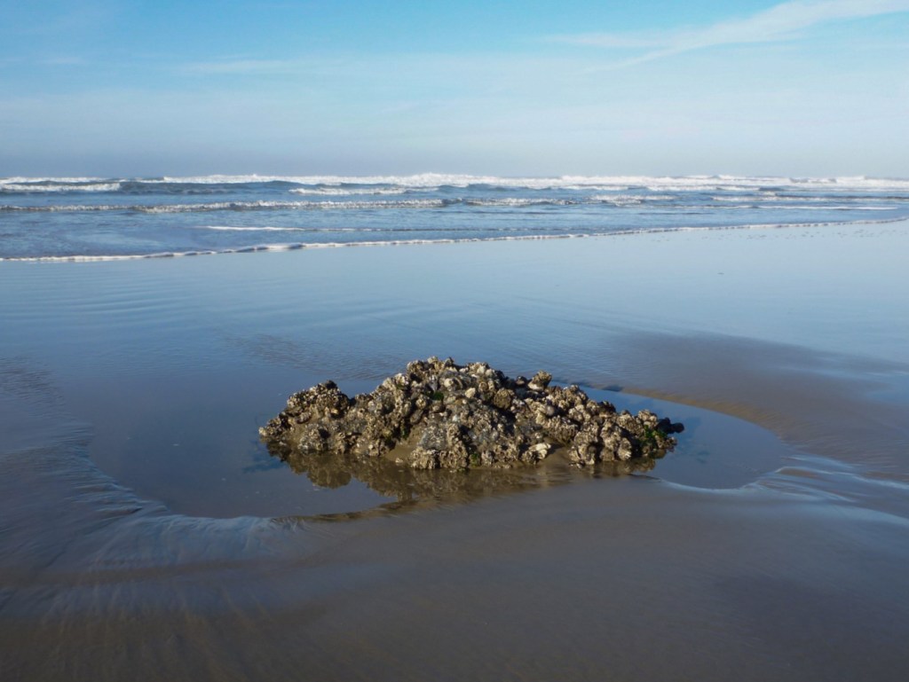 Beachscape with the tip of a rock barely emerging from intertidal sand. Surf zone in the background. mostly clear sky with offshore haze. 