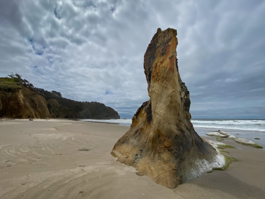A strange sandstone tower-like outcrop jutting out of beach sand; headland in the distance, surf zone to the right, cliffs to the left