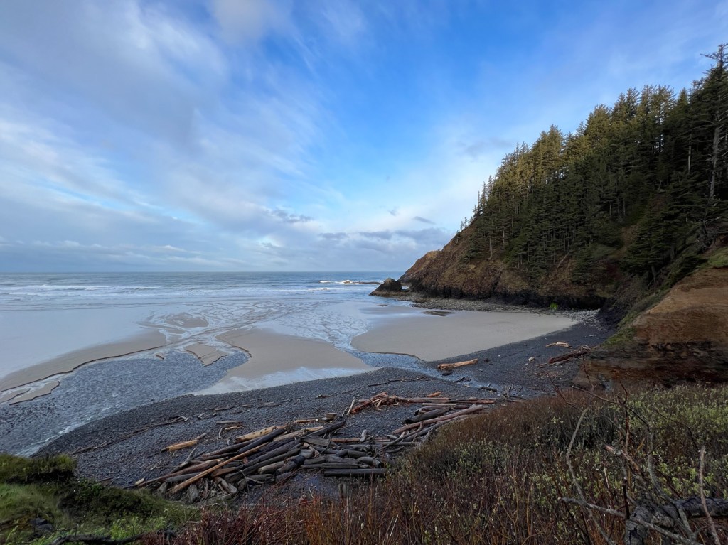 From above, a view out to sea, down across the cobbles, sand, sufzone, and beyond. Partly cloudy skies and a headland to the north.