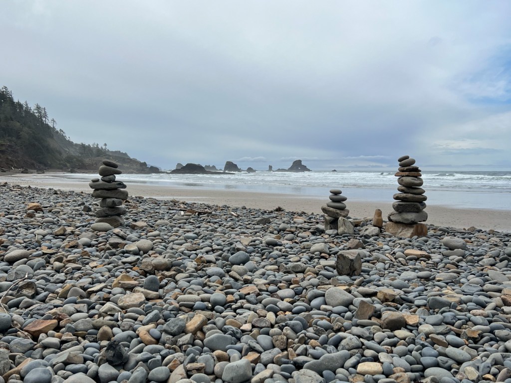 Three cobble cairns in the foreground, the view southwest over the cobble zone, onto sand, the out to the surf zone and beyond. A few offshore rocks in the distance and mostly cloudy skies.