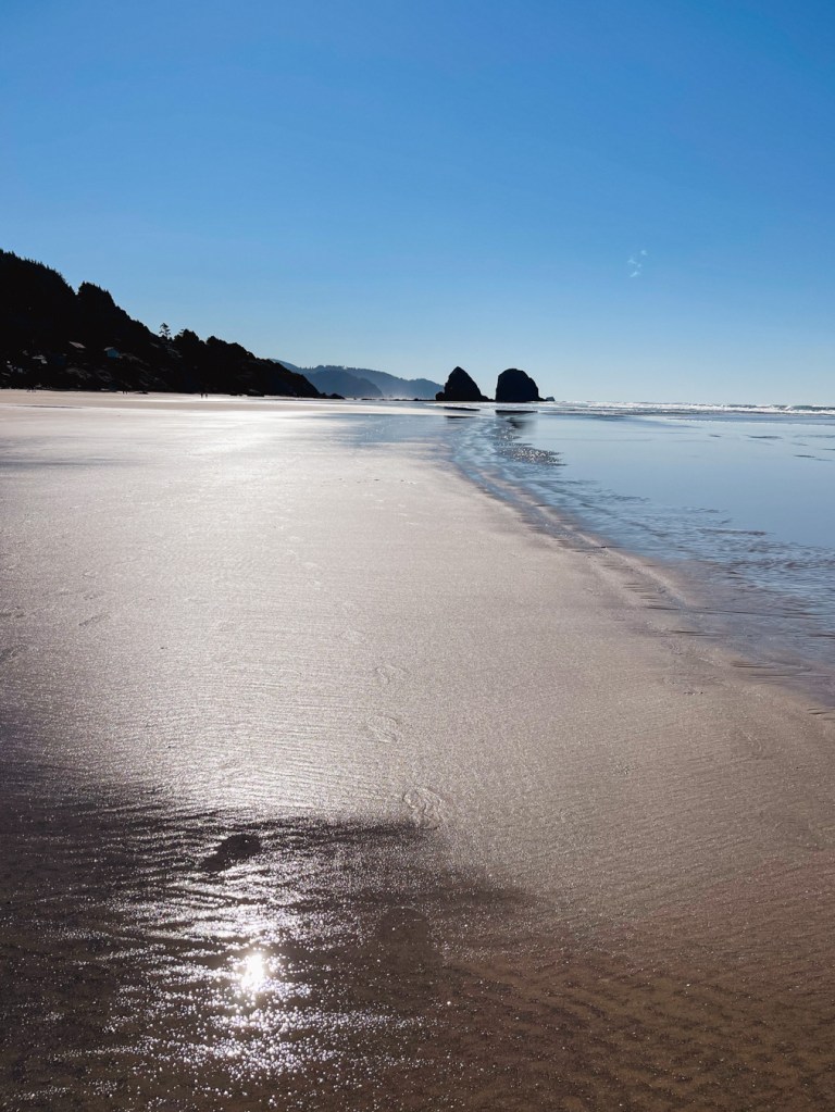 Long stretch of sandy beach with two sea stacks in the distance. Cloudless day with some sun gleam on the wet beach sand.