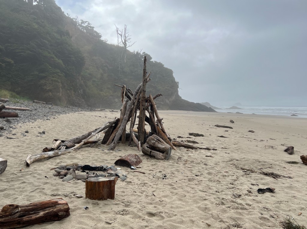 In the foreground some driftwood seats around a cobble campfire ring and beyond that, a teepee-like driftwood structure. In the left-hand distance, forested cliffs. To the right, the beach and way back, the surf zone. 