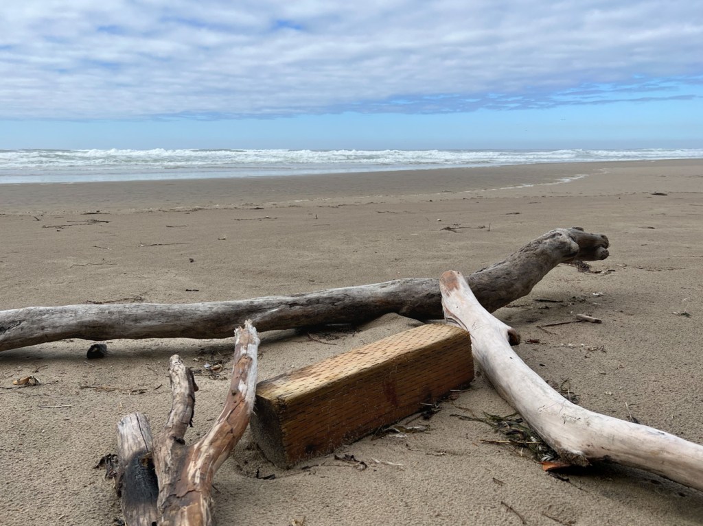 In the foreground, a section of pressure treated 6x6 and some drifted logs. Behind, the beach and surf zone. Out to sea, partly cloudy skies. 
