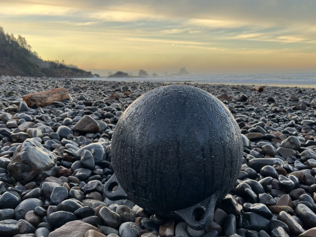 A two-lug float rests on cobbles shiny with indirect morning sunlight. Beach and the surf zone in the midground. The sun hasn't yet topped the low headland in the distance. Moody sky.