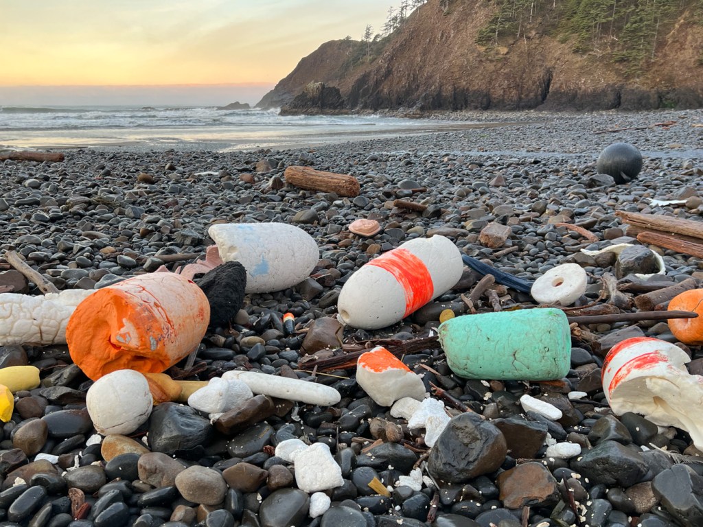 An accumulation of lost float and marker buoys in the cobbles. Surf zone and a headland in the distance. A kiss of sunrise in the sky.