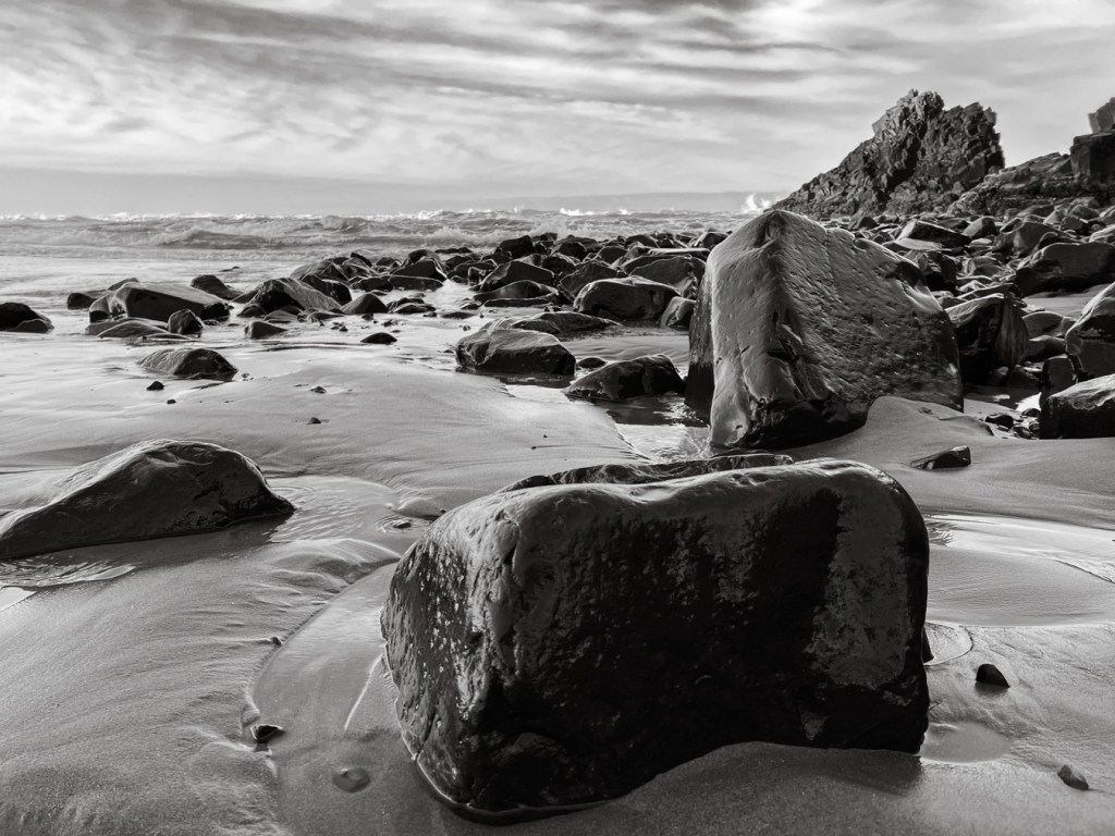 A boulder field and the incoming swash. In the distance, the surf zone and beyond. Partly cloudy sky.