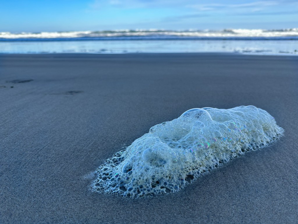 A small patch of sea foam rests on the sand. In the distance, the beach, mirror, and surf zone.