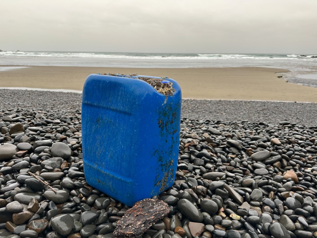 Plastic water or fuel container up on the cobble shelf. Beach and the surf zone in the distance. Cloudy sky.