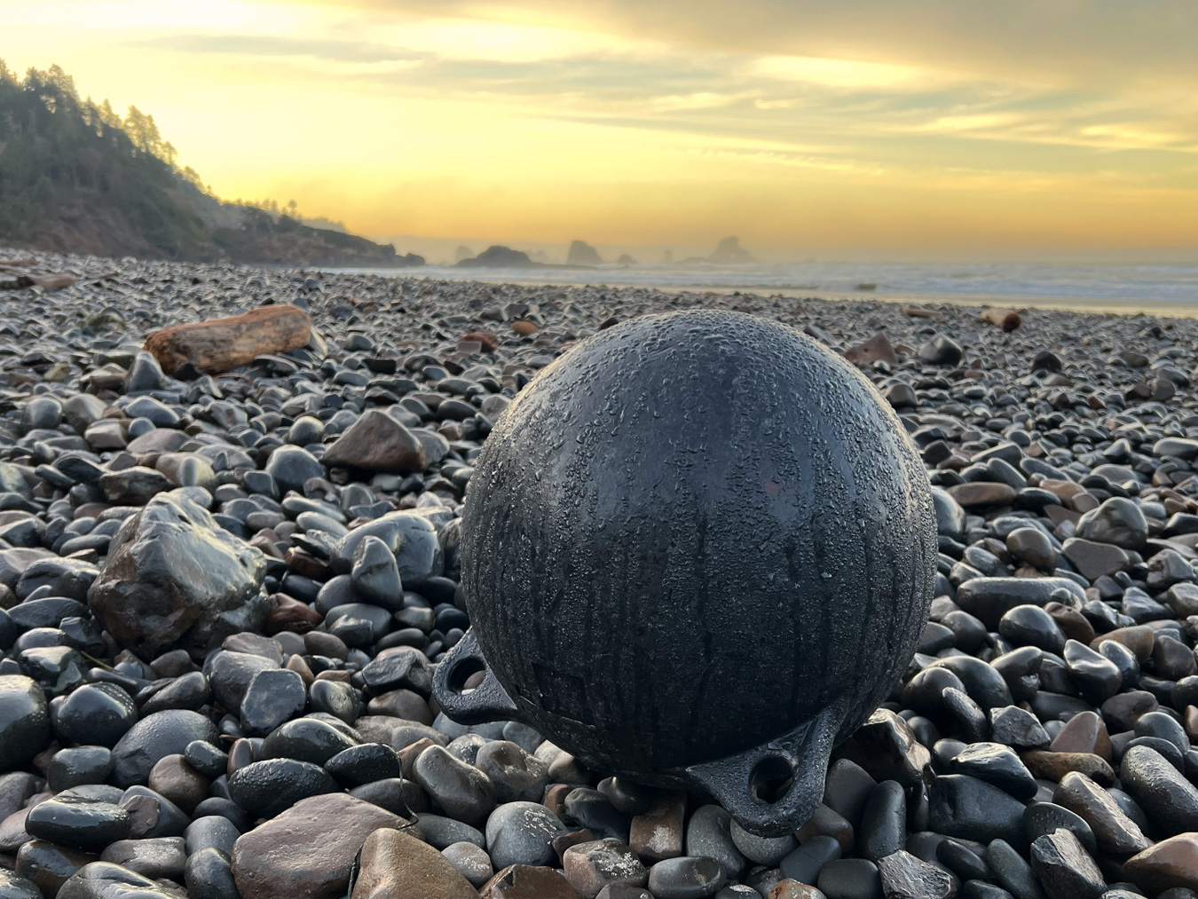 A lost buoy at rest in the cobbles. Surf zone and some offshore rocks in the distance.