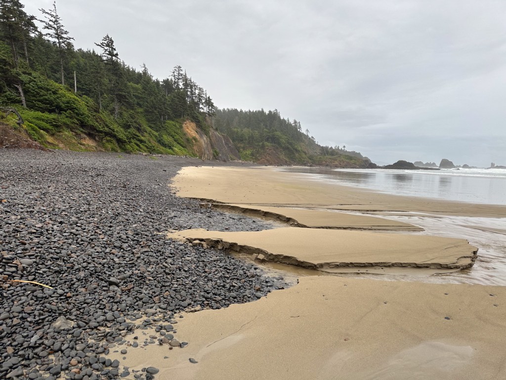 Seascape featuring the cobble szone, beach and surf zone. In the distance, crested slopes and offshore rocks.