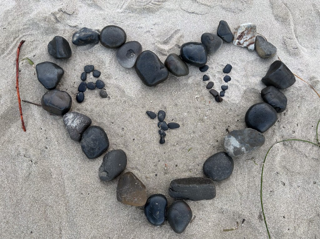 A cobblestone heart with "A + V" in pebbles on the sand. The highest swash has run up about halfway over the heart, slightly moving one of the pebbles in "+".