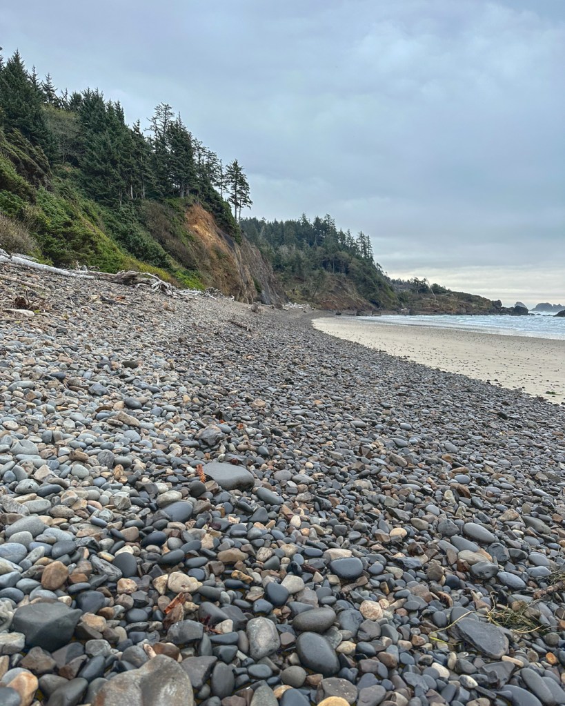 Beachscape with wet cobbles from the previous high tide and dry cobbles above the reach of tide. On the right, a headland; on the left, beach and the surf zone. Cloudy sky.