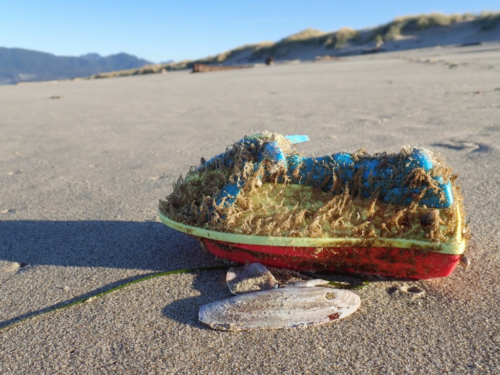 A heavily encrusted toy jet ski rest on the sand against a razor clam shell. Back beach and foredune in the background. Blue sky.