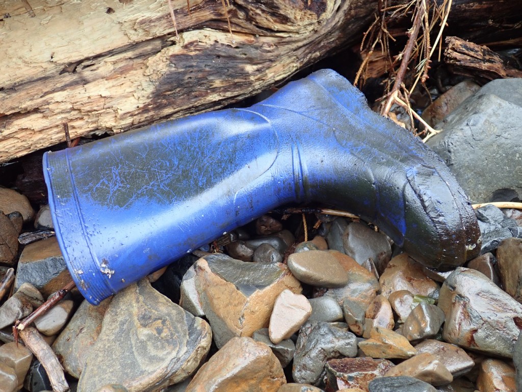 A blue rubber boot rests against driftwood on the cobbles.