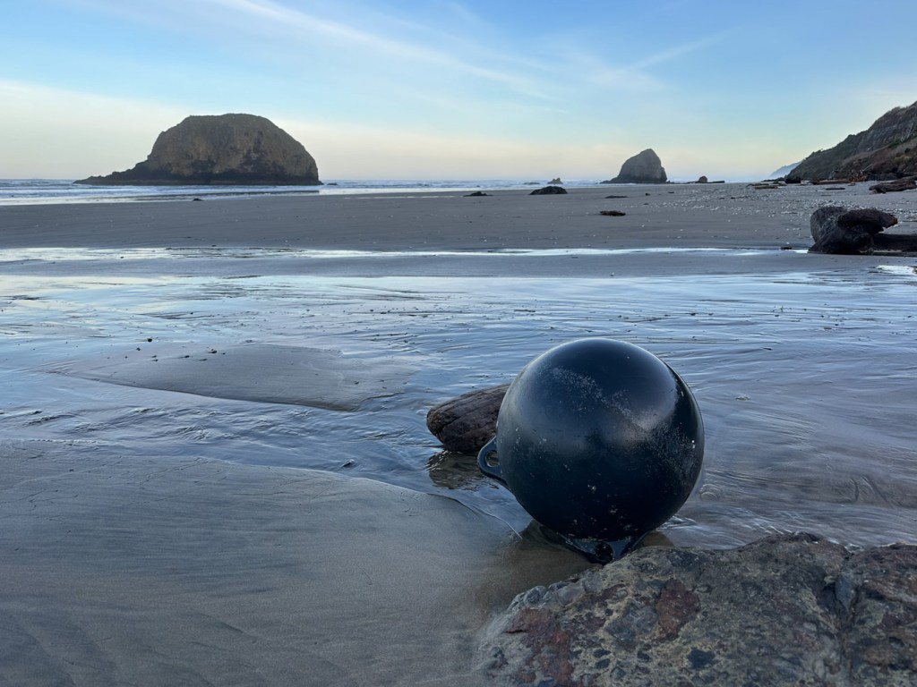 Kind of a seascape with a couple distant stacks, but in the foreground a two-lug round float rests on the beach. Mostly clear sky.