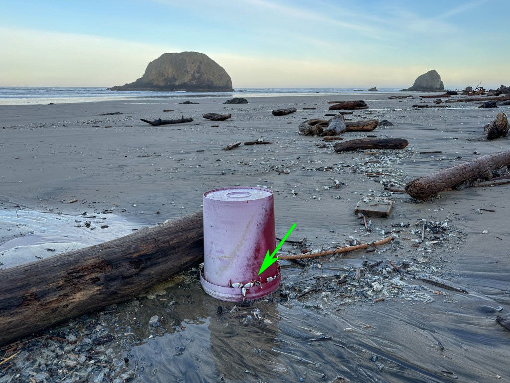 Kind of a seascape with a couple distant stacks, but in the foreground an empty bucket rests upside-down on the beach. Mostly clear sky.