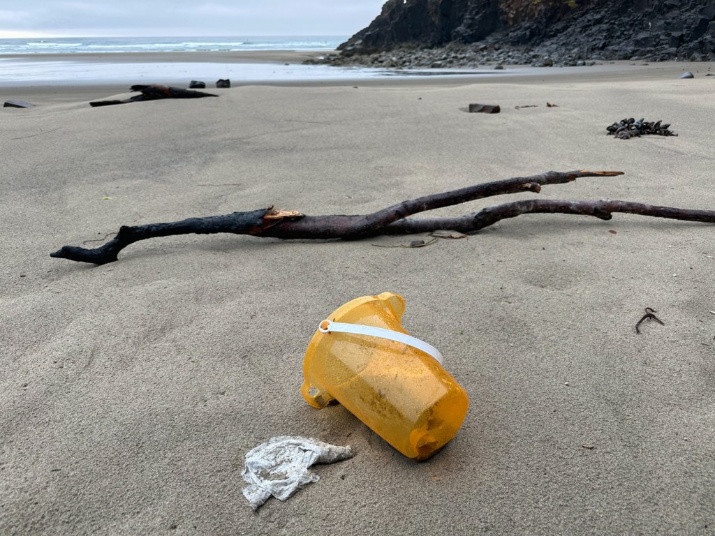 A child's plastic beach pail, lost of left behind, dominates the foreground in a beach scene. Rocky shore to the right; surf zone and beyond to the left. Cloudy sky.