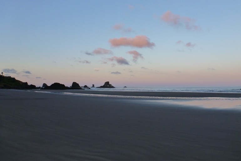Open dawn beachscape. In the foreground, the beach. Surf zone and a few rocks and the lowest slope of a headland. Mostly clear sky with a few small fluffy clouds. Color on the horizon.