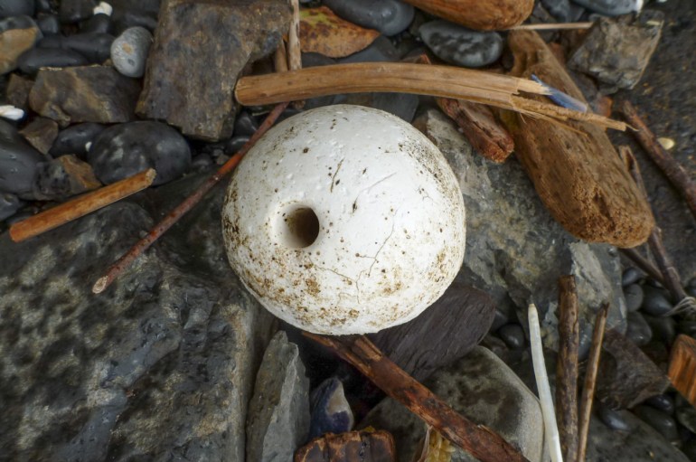 A lost and drifted center-hole trawl float rests among small driftwood on the cobbles.