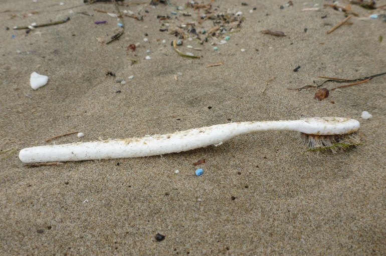A lost and drifted toothbrush rests on the sand among some tiny plastic fragments.
