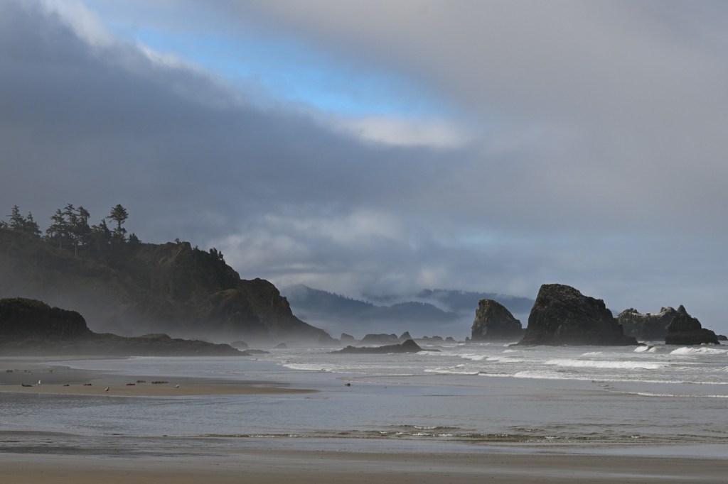 A seascape showing headlands and offshore rocks. Beach and the surf zone in the foreground. Mostly cloudy sky.