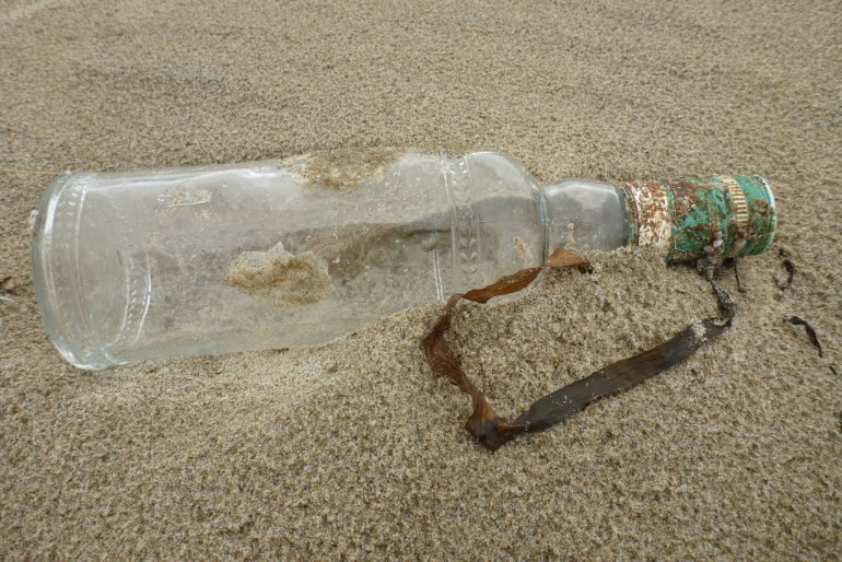 An empty and drifted Russian liquor bottle rests on the sand. The bottle is adorned with wheat heads. The label has a drawing of a bear and some wheat heads with the Russian words Sily Prirody, a Russian vodka brand, which translates to Forces of Nature or Powers of Nature.