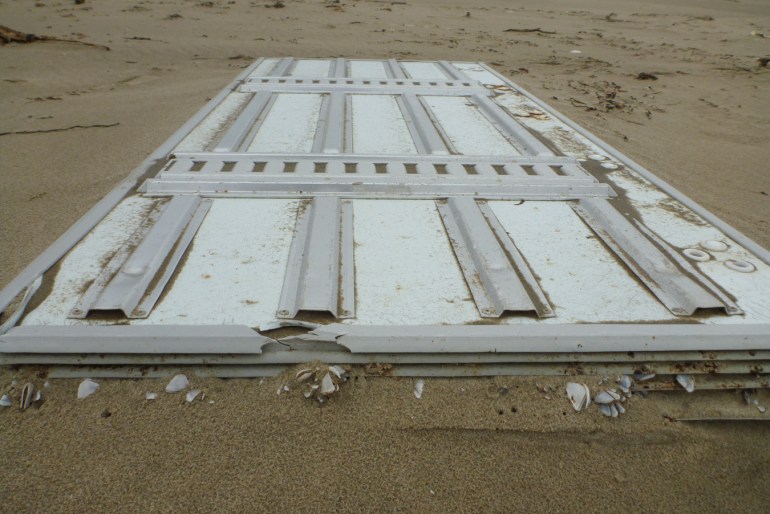 A large drifted insulated panel with a few pelagic barnacles attached rests on the sand.