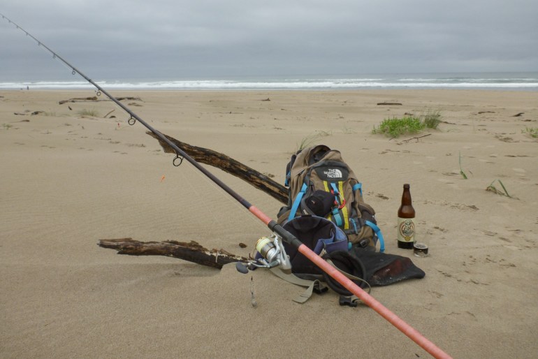 Backpack and surf fishing gear propped up on a piece of driftwood. Bottle of beer sitting on the sand, Beach and surf zone in the distance. Cloudy sky.