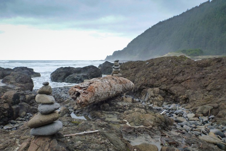 A pair of cairns and a big drifted log up on the rocks. The sea and a headland in the distance.