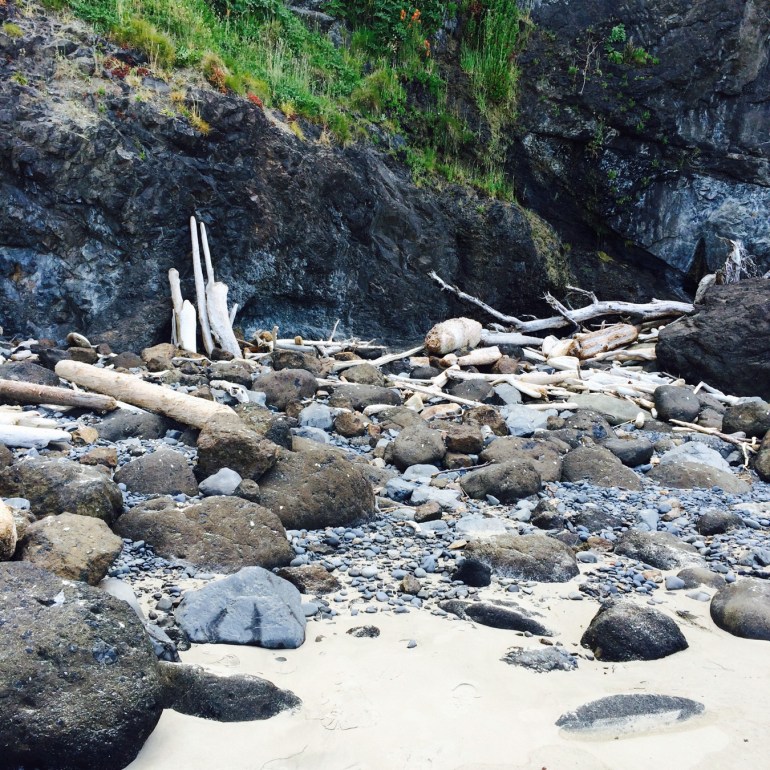 A beach scene with sand, cobbles, boulders, and driftwood up against the base of the cliff.
