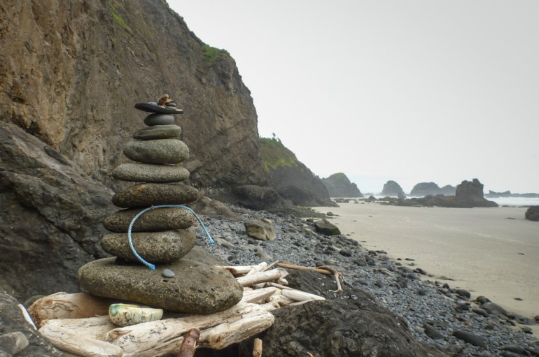 An attractive rock cairn built on a bed of small driftwood. Cobbles, beach and intertidal and offshore rocks in the distance. Hazy sky.