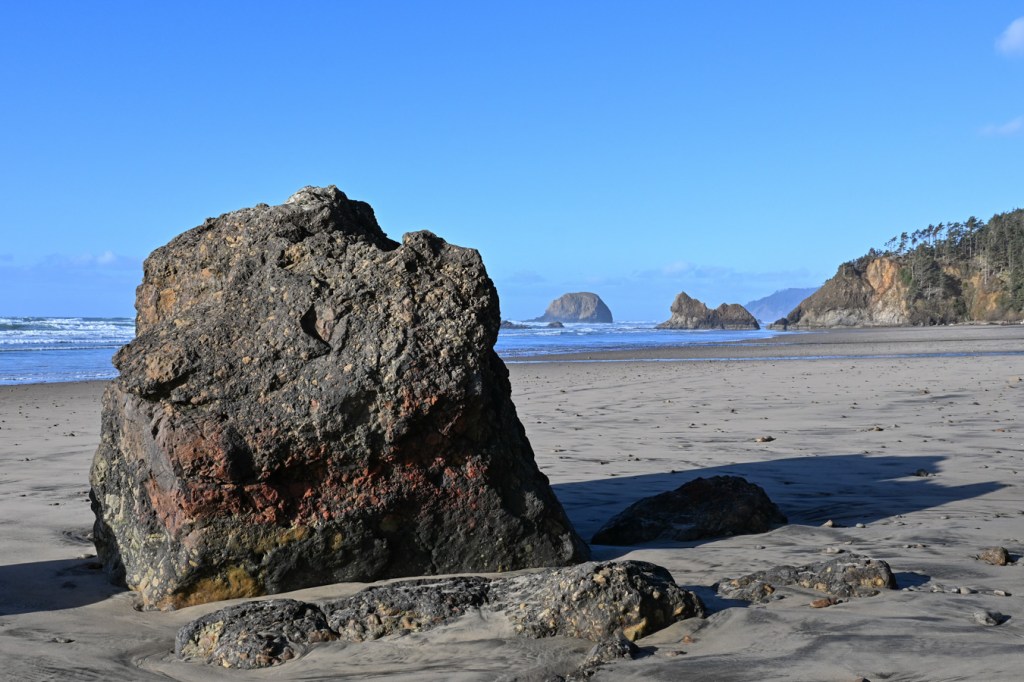 A seascape featuring the beach, jutting rocks, sea stacks, and headlands. Clear blue sky, with a few thin clouds in the distance.