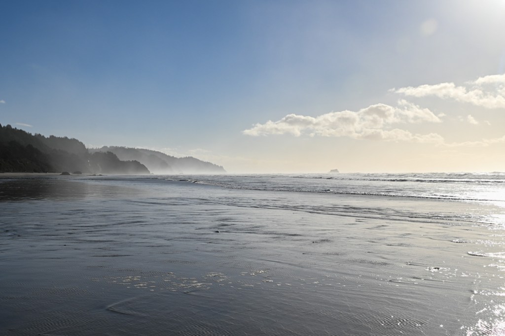 A beachscape with a series of headlands stretching off to the south. Blue sky with a few small puffy clouds.