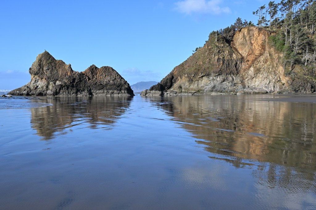 A seascape featuring a headland called Humbug Point and a sea stack known as The Lion set off against a swash-zone sheen in the foreground and mostly clear blue sky.