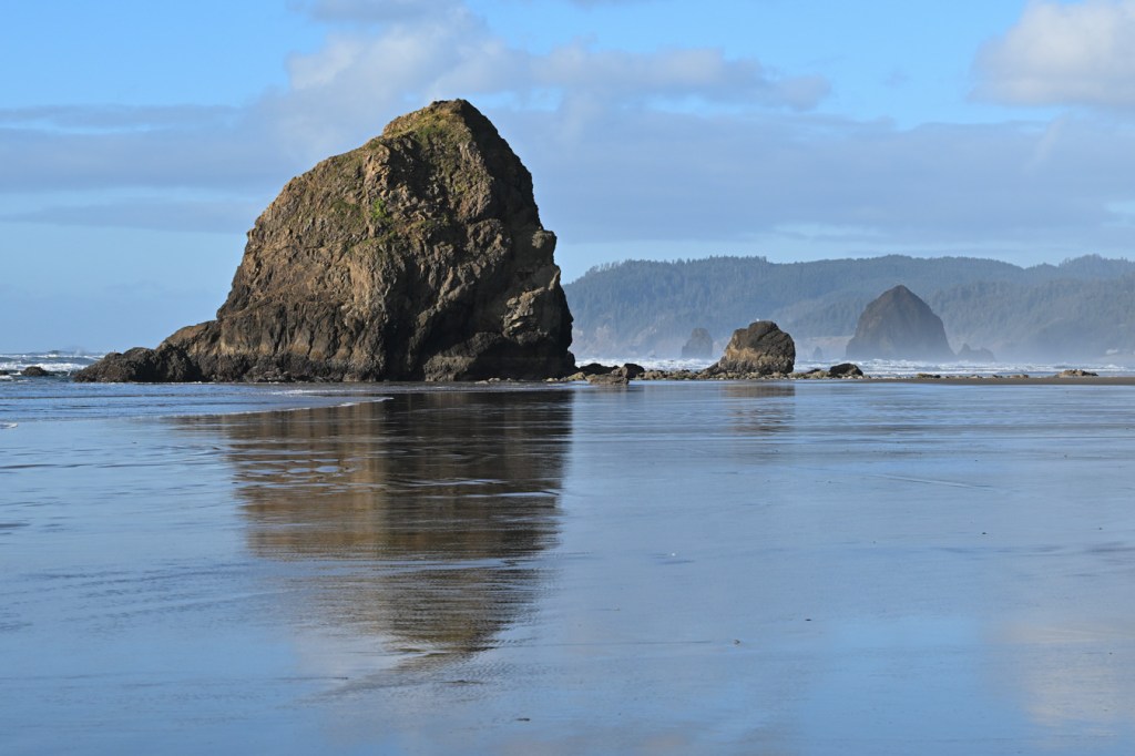 A seascape featuring iconic northern Oregon sea stacks set off against a swash-zone sheen in the foreground and a distant headland. Mostly blue sky with a few clouds.