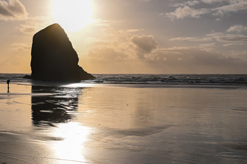 A golden-hour seascape with the sun at play above and behind a sea stack. A swash-zone mirror broken by reflections in the foreground. Partly cloudy sky.