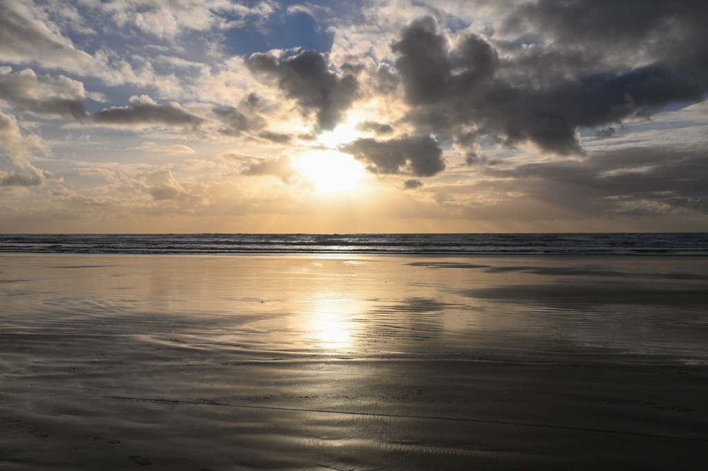 A golden-hour beachscape sunset looking straight into the sun. A swash-zone mirror in the foreground and quite a few clouds in the sky.