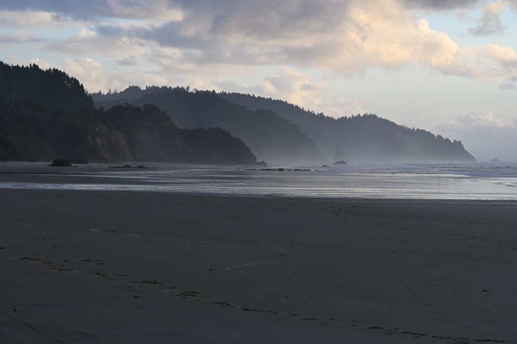 A late-afternoon seascape featuring a long sand beach and headlands stretching off into the distance. Partly cloudy sky.