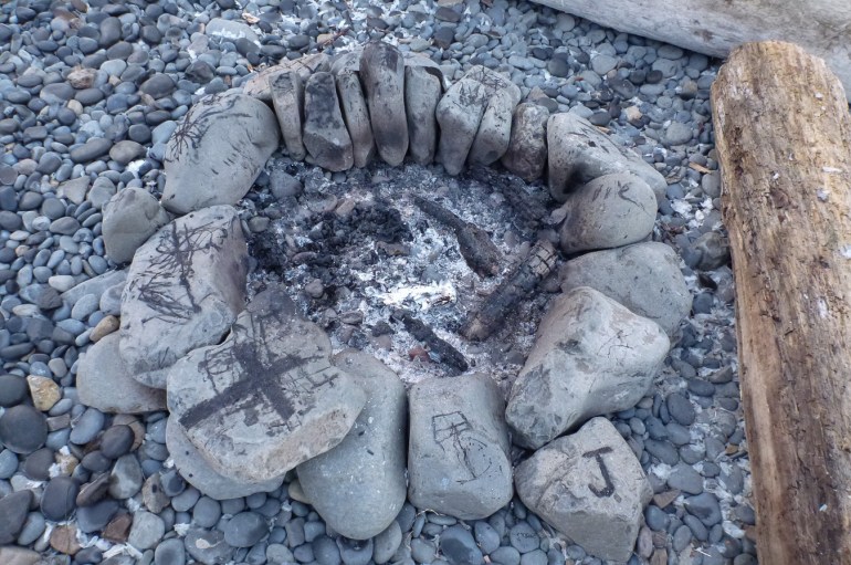 A campfire ring of large smooth rocks rests in, and blends in with, the cobbles. Driftwood "benches” are also visible at the scene's periphery.