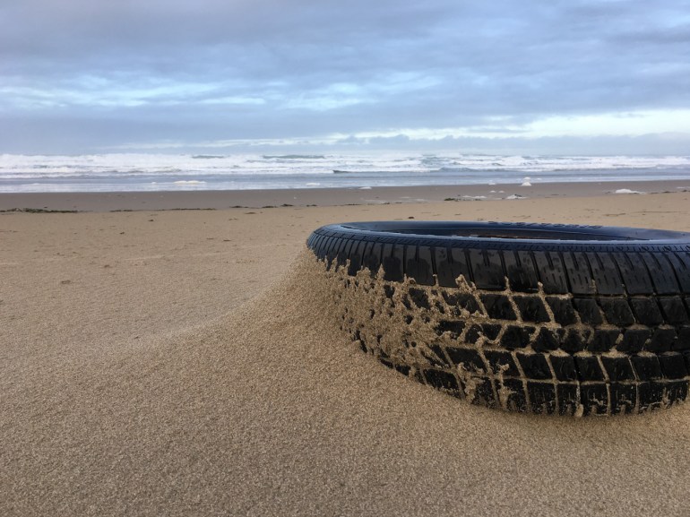 A drifted tire rests on windblown sand. Surf zone in the distance. Mostly cloudy sky.