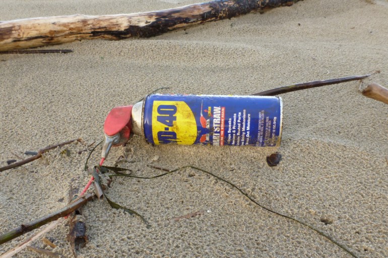 A WD-49 can rests among driftwood on rain-dimpled sand.
